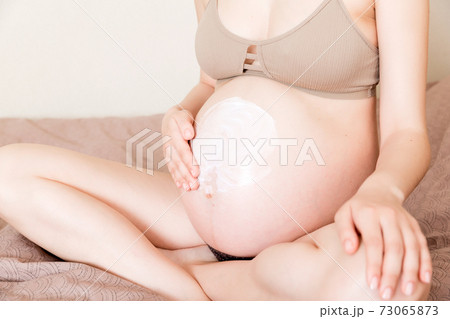 a pregnant girl sits at home on the bed and smears an anti-stretch mark cream on her stomach. Pregnancy, motherhood, preparation and expectation concept 73065873