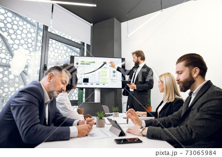 Business concept. Handsome bearded male team leader having a presentation on digital whiteboard, showing marketing and sales report of company to his multiracial colleagues 73065984