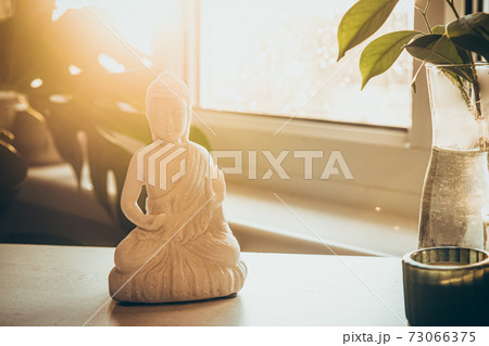 Decorative white Buddha statuette figure with candles standing on the tray near the window with a green monstera plant and bright sun light on the background. Meditation, relax ritual. Selective focus Decorative white Buddha statuette figure with candles standing on the tray near the window with a green monstera plant and bright sun light on the background. Meditation, relax ritual. Selective focus 73066375