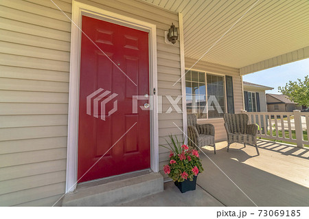 Shiny red wooden front door of a home with wicker chairs on the sunlit porch 73069185