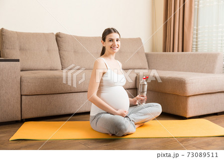 Pregnant young woman with bottle of water while sitting in lotus position on yoga mat. Maintaining water balance at coronavirus time Pregnant young woman with bottle of water while sitting in lotus position on yoga mat. Maintaining water balance at coronavirus time 73089511