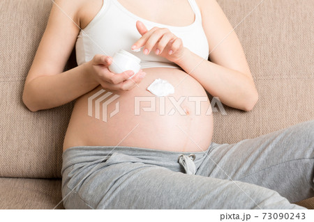 a pregnant girl sits at home on the bed and smears smile an anti-stretch mark cream on her stomach. Pregnancy, motherhood, preparation and expectation concept 73090243
