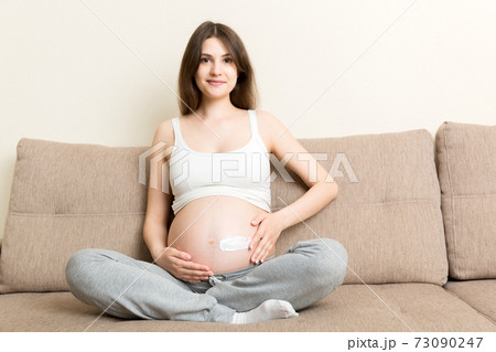 a pregnant girl sits at home on the bed and smears an anti-stretch mark cream on her stomach. Pregnancy, motherhood, preparation and expectation concept a pregnant girl sits at home on the bed and smears an anti-stretch mark cream on her stomach. Pregnancy, motherhood, preparation and expectation concept 73090247