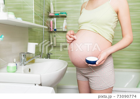 Young pregnant woman applying anti-stretch marks cream to the abdomen in the morning in the bathroom. Pregnancy, maternity and bodycare 73090774