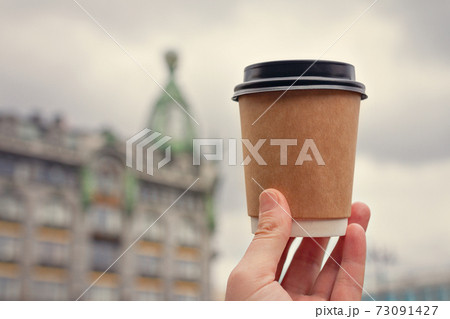 paper cup of natural coffee in woman's palm on background of Nevsky Prospect in St. Petersburg and cloudy sky 73091427