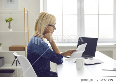 Serious senior woman sitting at her desk at home and reading notification letter Serious senior woman sitting at her desk at home and reading notification letter 73097349