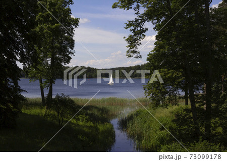 Beautiful landscape - green trees and meadows, a white boat sails in the distance of the lake. Lithuania, near Trakai 73099178