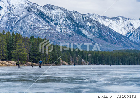 Tourists doing ice-skating in Johnson Lake frozen water surface in winter time. Snow-covered mountain in the background. Banff National Park, Canadian Rockies, Alberta, Canada. 73100183