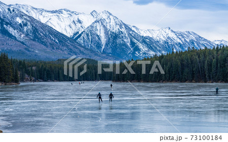 Tourists doing ice-skating in Johnson Lake frozen water surface in winter time. Snow-covered mountain in the background. Banff National Park, Canadian Rockies, Alberta, Canada. Tourists doing ice-skating in Johnson Lake frozen water surface in winter time. Snow-covered mountain in the background. Banff National Park, Canadian Rockies, Alberta, Canada. 73100184