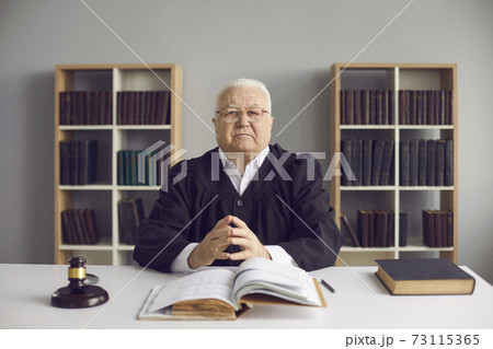 Wise mature judge sitting at desk with open Code of Law in his office or courtroom Wise mature judge sitting at desk with open Code of Law in his office or courtroom 73115365