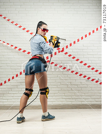 a slender woman in the image of a construction worker with a puncher against a white textured brick wall 73117019