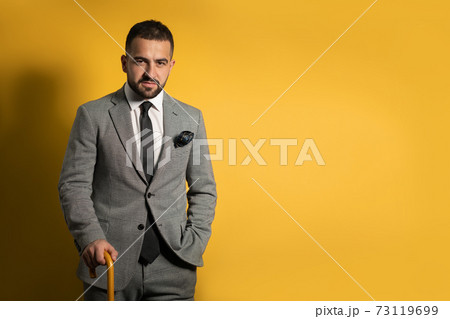 Old school trendy stylish handsome young man wearing grey suit with one hand lifted standing with cane or umbrella in his hand isolated on yellow background Old school trendy stylish handsome young man wearing grey suit with one hand lifted standing with cane or umbrella in his hand isolated on yellow background 73119699