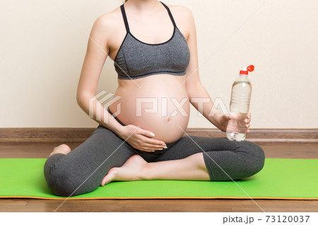 Pregnant young woman with bottle of water while sitting in lotus position on yoga mat. Maintaining water balance at coronavirus time 73120037