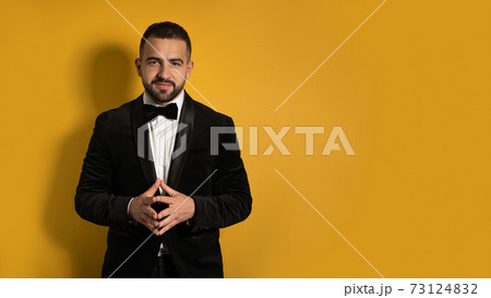 Handsome man in tuxedo standing fingers folded down smiling on camera, work profession lifestyle. Handsome young smiling caucasian man isolated on yellow background 73124832