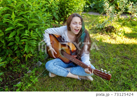 Young woman sitting in grass and playing guitar Young woman sitting in grass and playing guitar 73125424