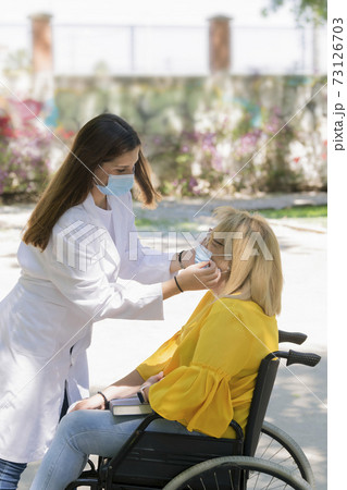 Young female caregiver carefully puts a surgical mask on a mature woman using a wheelchair. safety concept. Young female caregiver carefully puts a surgical mask on a mature woman using a wheelchair. safety concept. 73126703