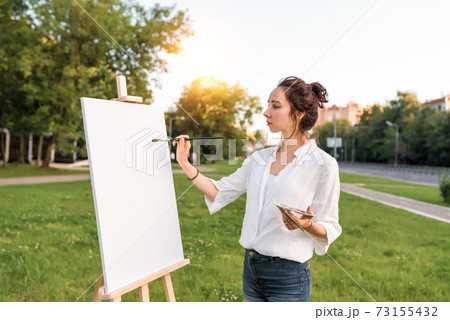 woman paints picture landscape, summer in city on lawn, girl artist white shirt, creating creativity artistic mood. Blank white canvas getting started. Highway background. Brush color palette paints. 73155432