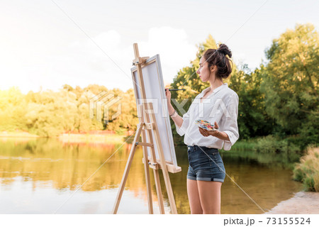 girl artist, woman paints picture of landscape, summer lake river pond, white shirt denim shorts, creating creativity artistic. Blank white canvas. Background trees water. Brush color palette paints. 73155524