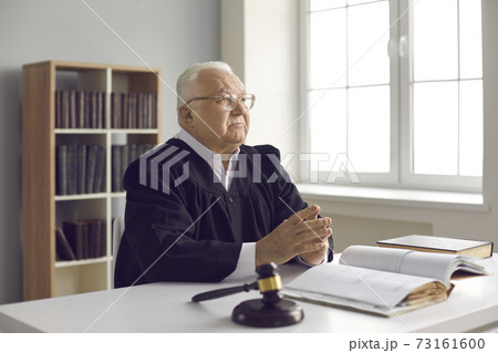 Serious mature judge sitting at desk with open law book in his office or courtroom Serious mature judge sitting at desk with open law book in his office or courtroom 73161600