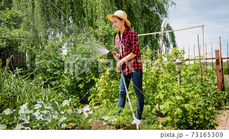 Smiling young girl holding water hose watering growing vegetables at backyard garden 73165309