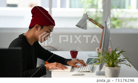 Side view of freelance graphic designer sitting at his workspace and looking at notebook while working on computer. 73176296