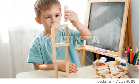 Happy smiling boy building tower with wooden blocks and bricks on desk at kindergarten classroom. Concept of smart children and home education during lockdown and self isolation. 73184089