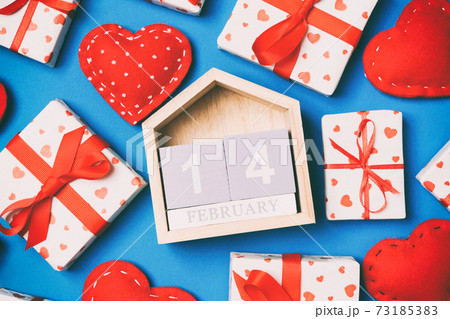 Composition of wooden calendar, holiday white gift boxes and red textile hearts on colorful background. The fourteenth of February. Valentine's day concept 73185383