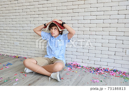 cute and smart little boy in blue shirt and glasses sitting with red books on his head with intended face and looking at something beside white wall background cute and smart little boy in blue shirt and glasses sitting with red books on his head with intended face and looking at something beside white wall background 73186813