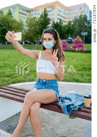 female woman in city summer in medical mask, sits on bench pictures smartphone, online recording on Internet, application phone, video call white topic casual clothes denim jacket, cup of coffee tea. 73187834
