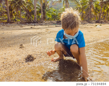 Cute little young blond boy with medical face mask on the beach playing with sand. Protecting from air pollution and virus infection as new normal lifestyle during the covid19 virus epidemic era Cute little young blond boy with medical face mask on the beach playing with sand. Protecting from air pollution and virus infection as new normal lifestyle during the covid19 virus epidemic era 73194894