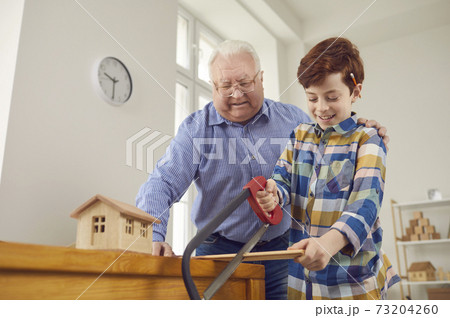 Smiling little grandson sawing a wooden plank while his grandfather is watching Smiling little grandson sawing a wooden plank while his grandfather is watching 73204260