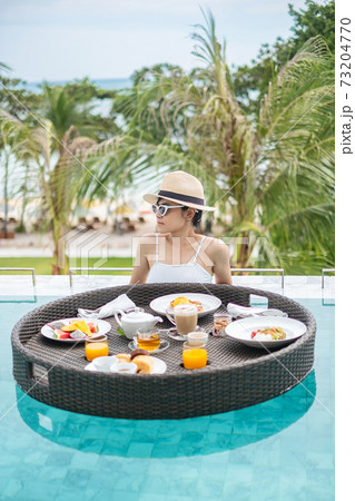 Happy woman in white swimsuit having Floating Breakfast tray in luxury pool hotel, young female with sunglasses enjoy in tropical resort. Relaxing, Exotic summer travel, holiday, vacation and weekend 73204770