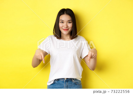 Beauty and fashion concept. Beautiful asian woman in white t-shirt pointing fingers down, demonstrate logo standing over yellow background 73222556