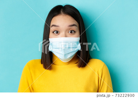 Covid-19, social distancing and pandemic concept. Close up of surprised asian woman in medical mask, raise eyebrows and look amazed at camera, blue background 73222930