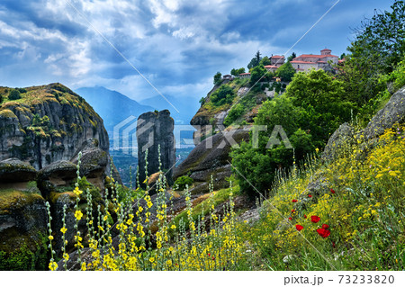 Typical view of Meteora, Greece, cliff monastery, sedimentary rocky pillars, yellow and poppy flowers. Sun rays fall perpendicular, clouds, mountains Typical view of Meteora, Greece, cliff monastery, sedimentary rocky pillars, yellow and poppy flowers. Sun rays fall perpendicular, clouds, mountains 73233820