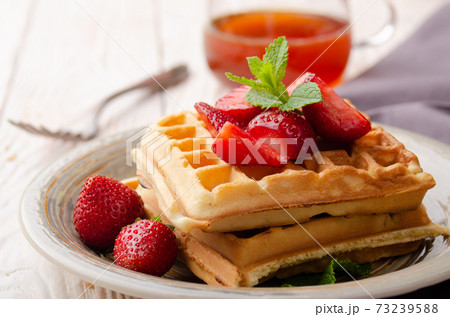 Closeup view at belgian waffles served with strawberries and mint leaf on white wooden kitchen table with syrup aside 73239588