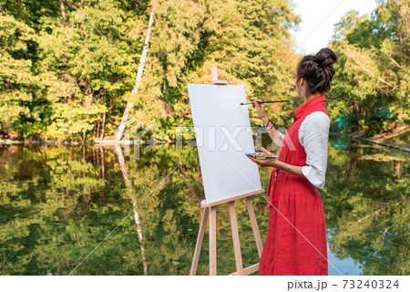free space for text, woman artist summer park near lake, river pond, draws a picture, white canvas. Creation creativity, art fantasy. Mirror reflection in water, brush colored paints girl. 73240324