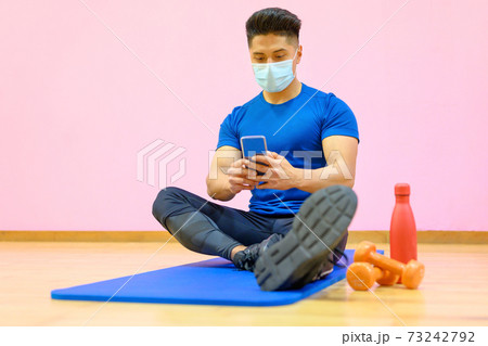 Young Latin man with protective face mask, relaxed, using mobile phone after workout in gym during 73242792