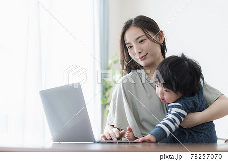 0歳の赤ちゃんを抱えたままパソコンを操作する若い母親 73257070
