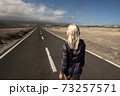 Blonde long hair girl viewed fromback walk on a long straight asphalt road - concept of freedom and independence - hope and future for next destination in life 73257571