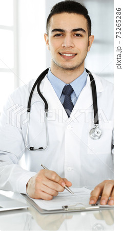 Arab doctor man is using laptop computer while filling up medication history records form at the glass desk in r clinic. Medicine concept Arab doctor man is using laptop computer while filling up medication history records form at the glass desk in r clinic. Medicine concept 73265286