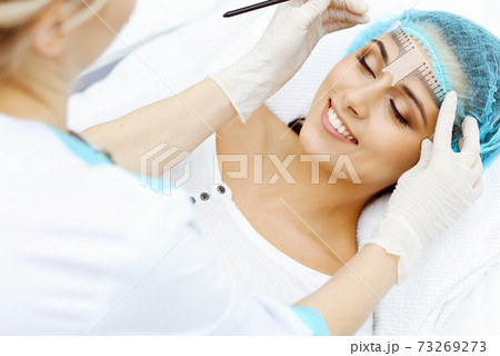 Professional beautician is going to correct eyebrow line at woman face using cosmetic ruler. Permanent brow makeup in beauty salon, closeup. Cosmetology treatment Professional beautician is going to correct eyebrow line at woman face using cosmetic ruler. Permanent brow makeup in beauty salon, closeup. Cosmetology treatment 73269273