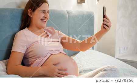 Smiling pregnant woman waiting for baby sitting in bed and calling friend or doctor on video call conference. Pregnant woman using smartphone 73270356