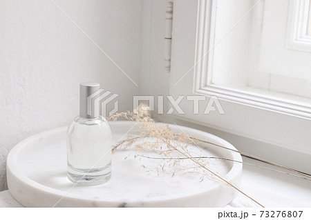 Feminine cosmetic still life scene. Glass bottle, flacon with perfume or eau de toilette and dry grass on white marble tray near window. Selective focus, blurred window sill background. 73276807