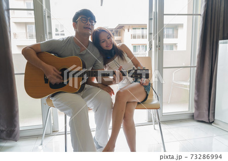 Romantic couple sit in the chair, play the guitar and listen to the music happily in the room. Romantic couple sit in the chair, play the guitar and listen to the music happily in the room. 73286994