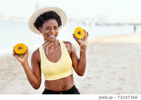 A young African girl in a summer straw hat is sitting on the beach, holding a papaya and smiling 73297366