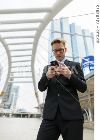 Young handsome blond businessman with eyeglasses using phone in the city outdoors Young handsome blond businessman with eyeglasses using phone in the city outdoors 73298835