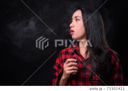 Young beautiful Asian woman vaping against black background Young beautiful Asian woman vaping against black background 73301421