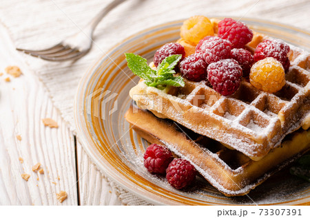 Belgian waffles served with raspberries and mint leaf dusted with powdered sugar on white wooden kitchen table 73307391