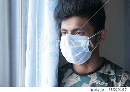 A young man with protective mask looking through window  73309167